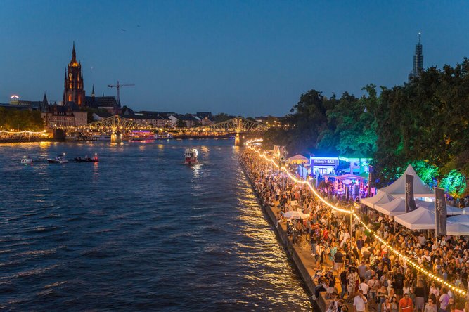 Crowds lining the Main River during Frankfurt’s Museumsuferfest, with festive lights, food stalls and the illuminated Kaiserdom in the background