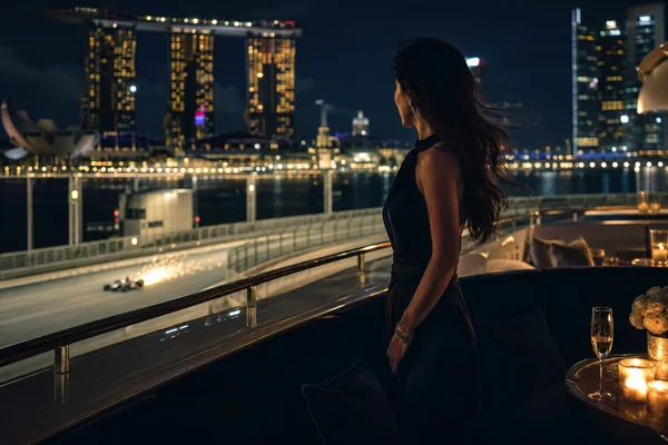 Singapore luxury yacht scene featuring an elegant woman in eveningwear overlooking Marina Bay Sands, the night skyline, champagne, candlelit seating, and a Formula 1 car on the illuminated circuit